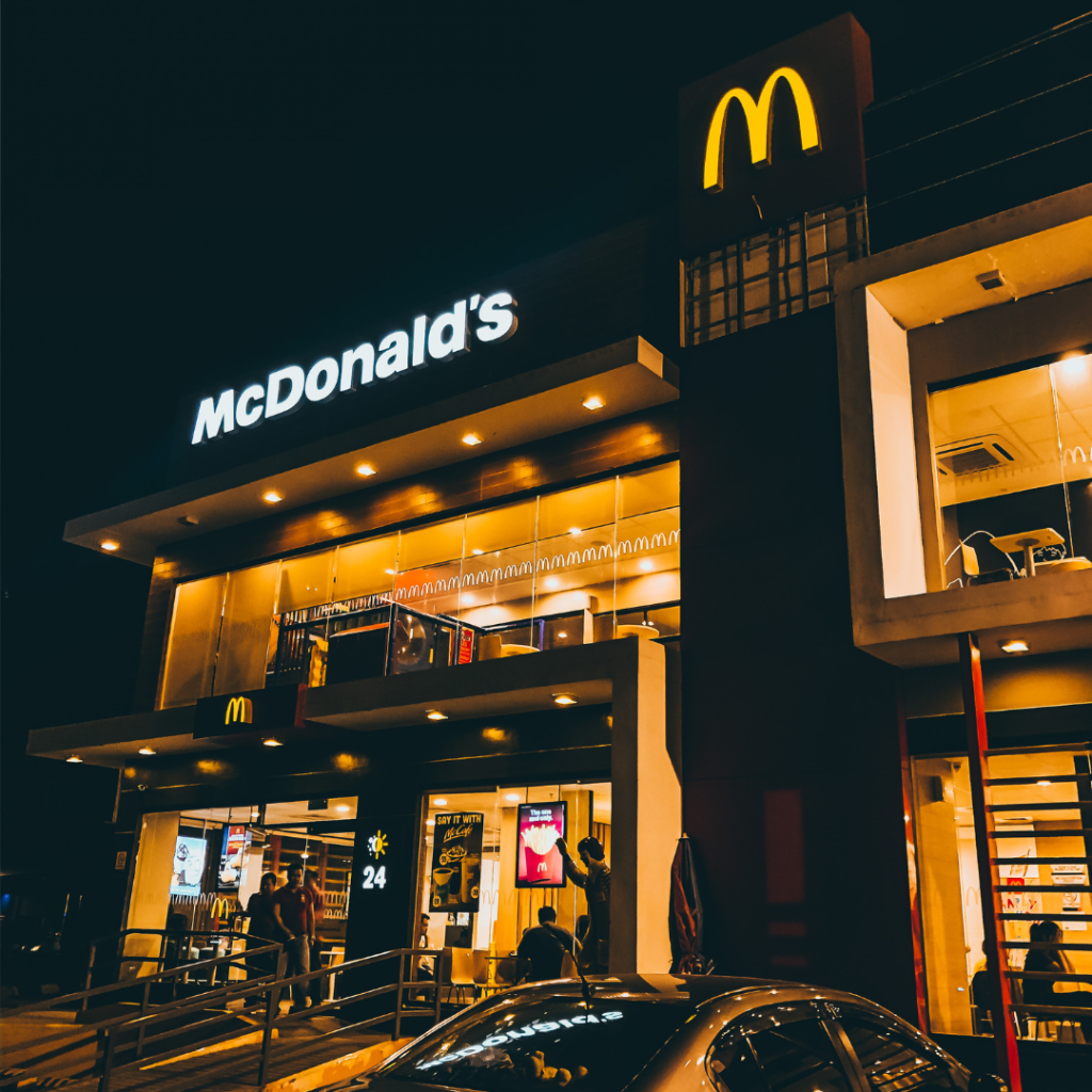 A imagem mostra uma fachada de um restaurante McDonald's à noite, com a iluminação amarela destacando as características modernas do prédio. O icônico logotipo do "M" dourado é visível no topo do edifício, assim como a marca "McDonald's" iluminada em branco. A entrada do restaurante possui grandes janelas de vidro que permitem ver o interior bem iluminado, com algumas pessoas visíveis dentro. O design é contemporâneo, com linhas retas e uma aparência sofisticada. Um carro está estacionado em frente ao restaurante, e o ambiente noturno cria uma atmosfera acolhedora e convidativa.